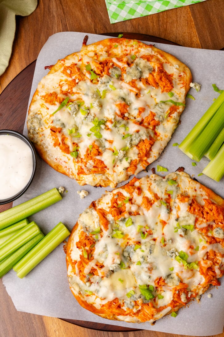 Flatbread topped with buffalo chicken, blue cheese, celery and mozzarella then baked on a wooden cutting board topped with parchment paper.