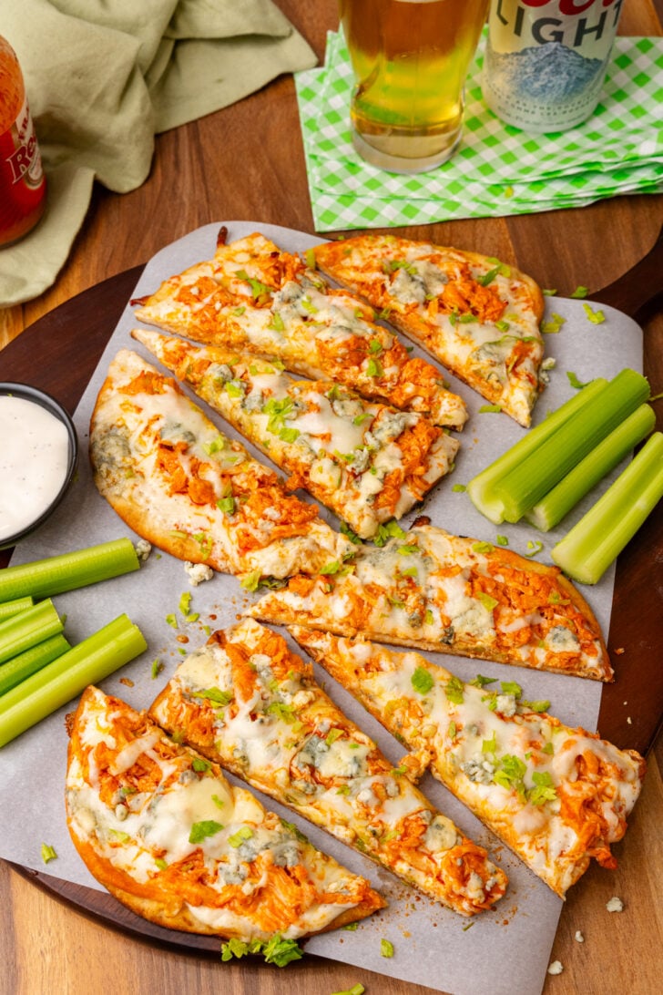 Flatbread topped with buffalo chicken, blue cheese, celery and mozzarella then baked, cut into triangles on a wooden cutting board topped with parchment paper.