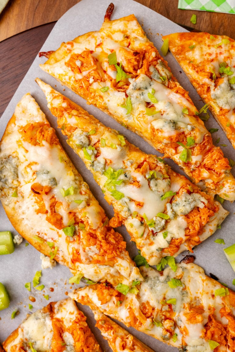 Flatbread topped with buffalo chicken, blue cheese, celery and mozzarella then baked, cut into triangles on a wooden cutting board topped with parchment paper.