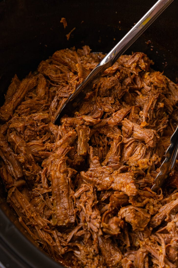 Black crock pot filled with cooked bbq beef roast shredded and served with metal thongs.