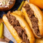 BBQ beef cooked in the crock pot served on small french rolls with slaw and pickle spears resting on parchment paper.