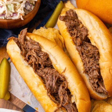 BBQ beef cooked in the crock pot served on small french rolls with slaw and pickle spears resting on parchment paper.