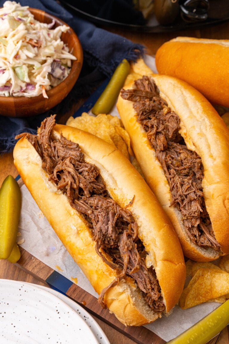 BBQ beef cooked in the crock pot served on small french rolls with slaw and pickle spears resting on parchment paper.