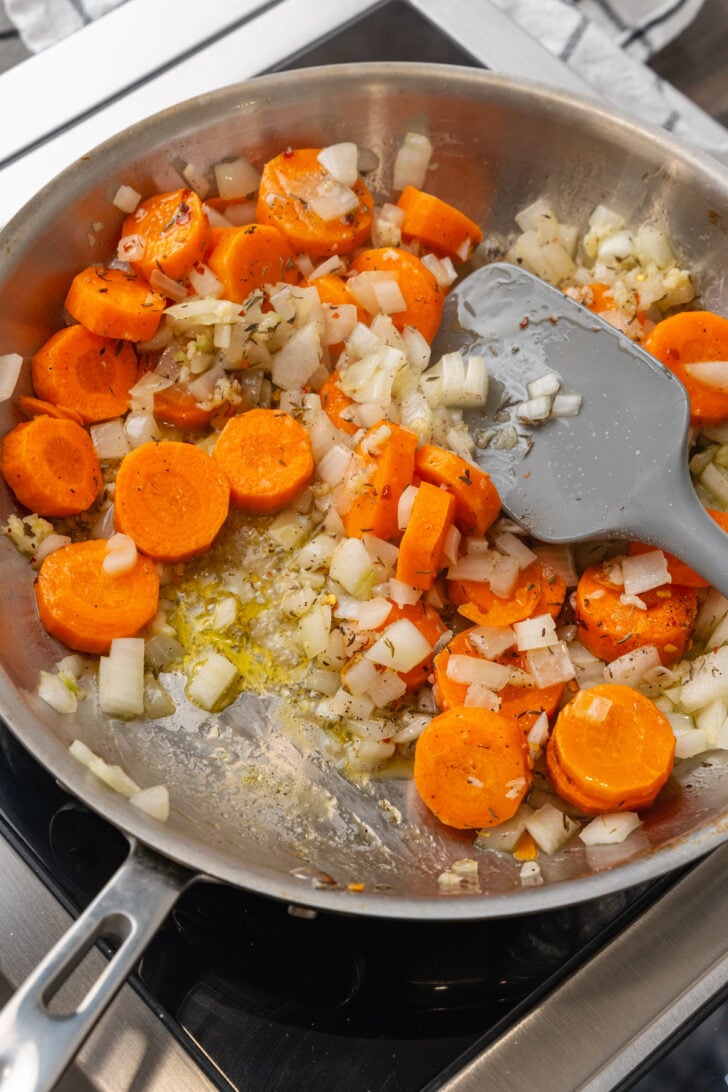 Metal skillet with peeled, sliced carrots, chopped onions and minced garlic cooked in olive oil with a spatula.