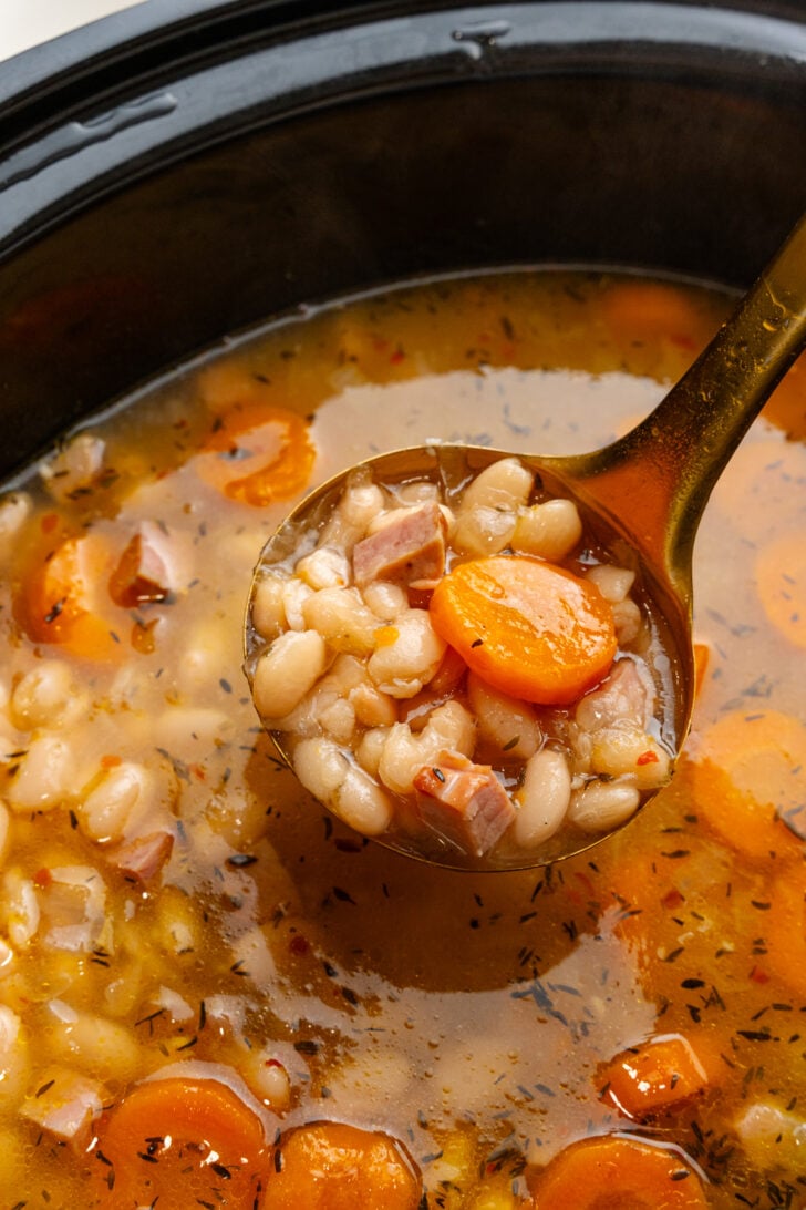 Gold ladle with white bean and ham soup in the bowl of a crock pot.