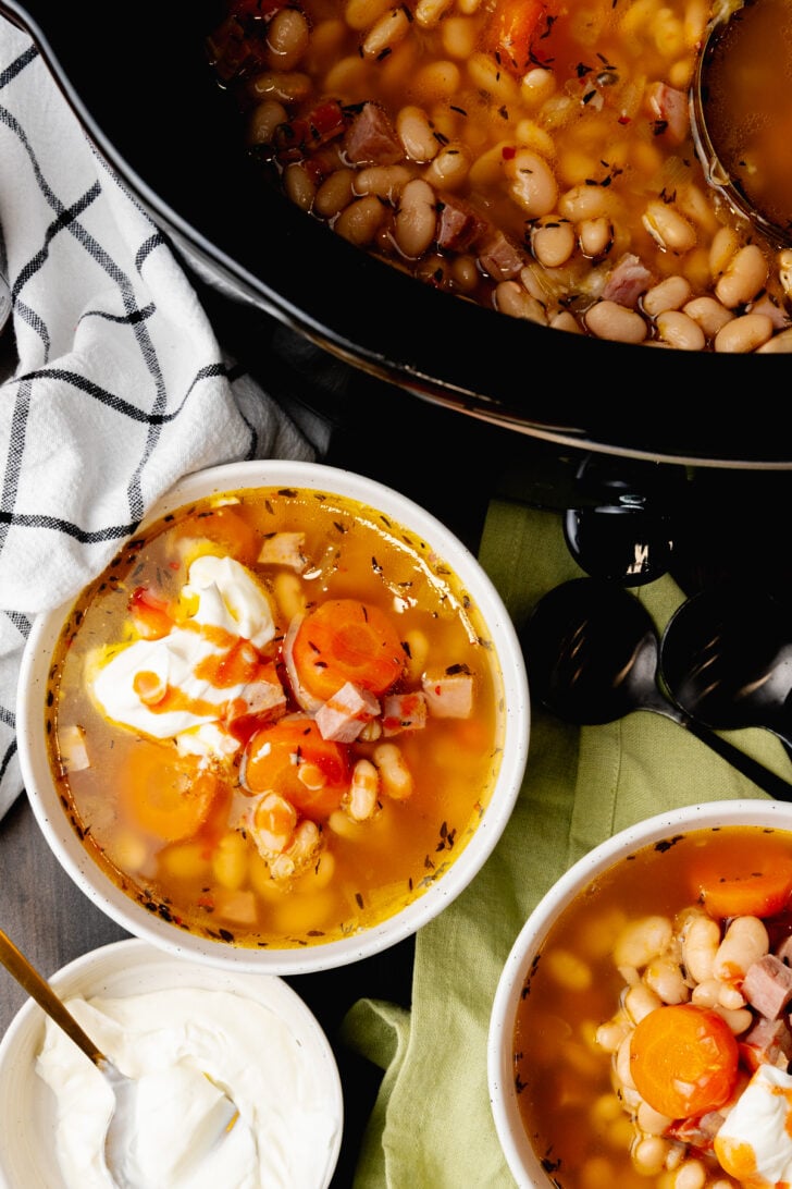 Two hite bowls filled with ham and white bean soup with a black spoon, topped with sour cream and hot sauce and a whole black crock pot full of soup.