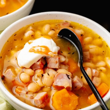 White bowl with ham and white bean soup with a black spoon, topped with sour cream and hot sauce.