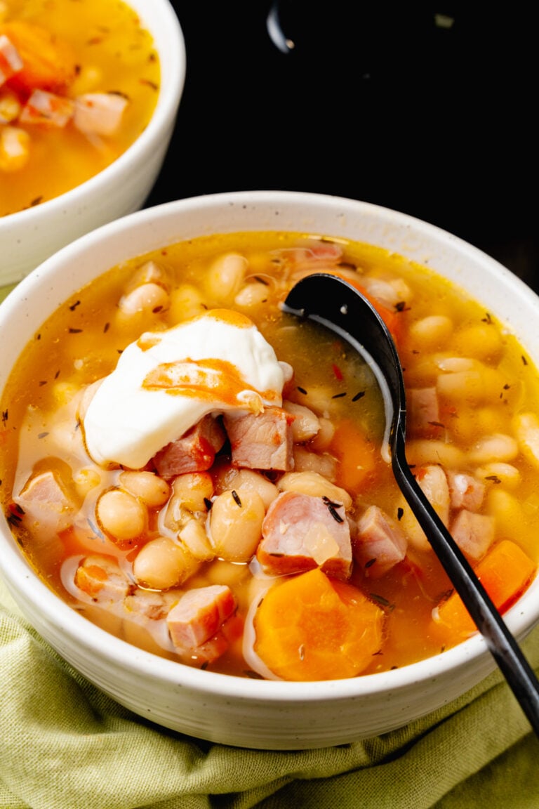 White bowl with ham and white bean soup with a black spoon, topped with sour cream and hot sauce.