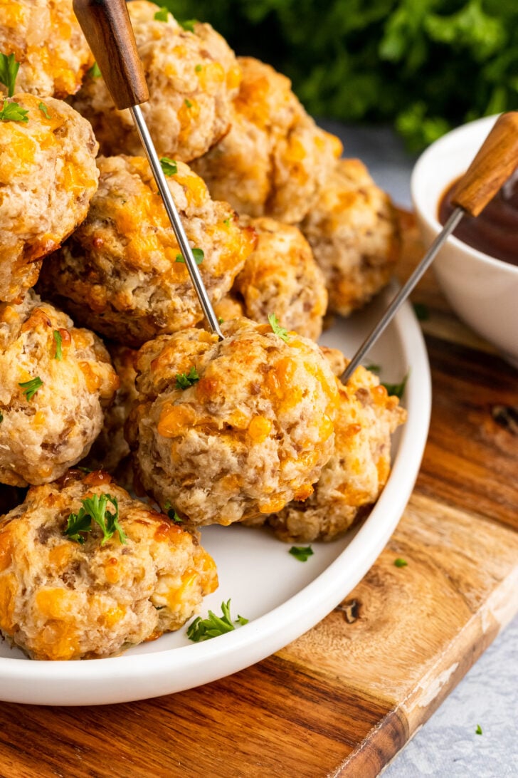Cream cheese sausage balls served with metal skewers on a white plate with fresh parsley resting on a wooden serving board.