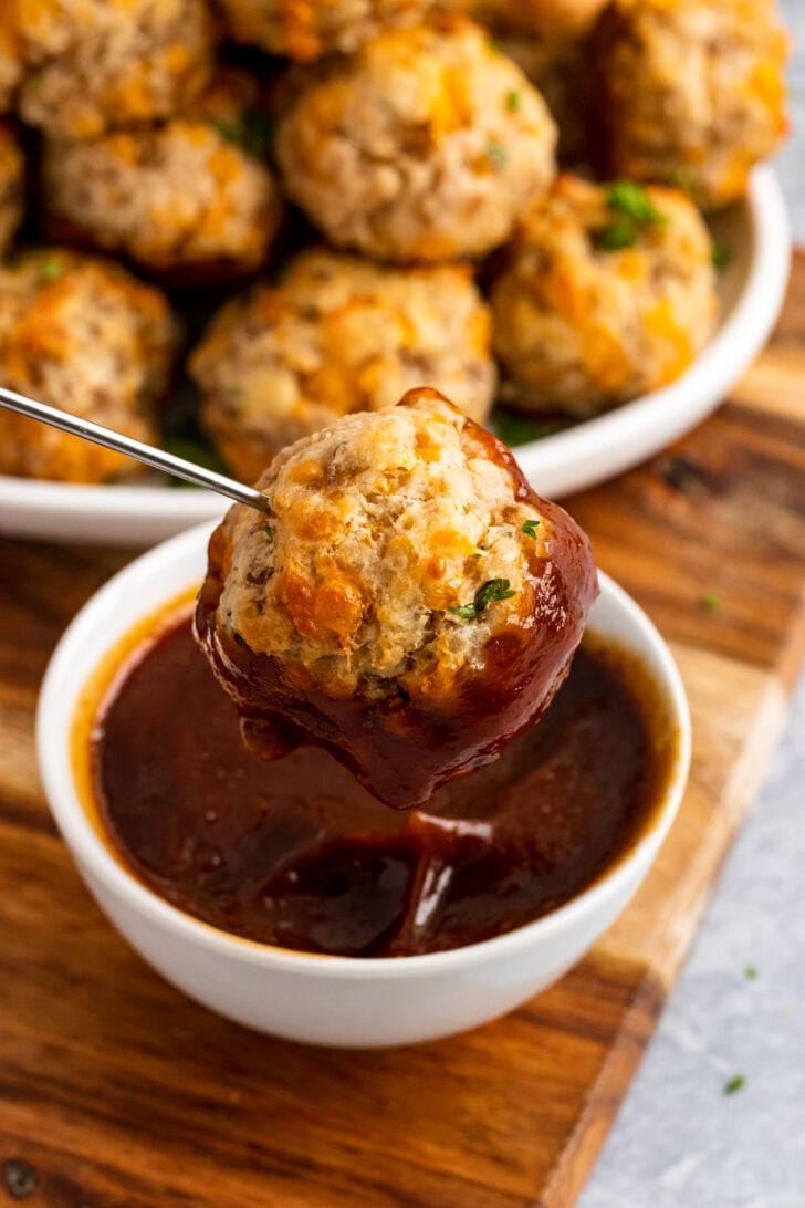 Cream cheese sausage ball on a metal skewer dipped into warm BBQ sauce in a small white bowl.