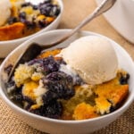 White bowl with blueberry cobbler and a scoop of vanilla ice cream resting on a tan placemat with silver forks.