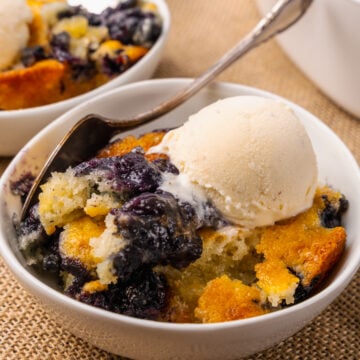 White bowl with blueberry cobbler and a scoop of vanilla ice cream resting on a tan placemat with silver forks.