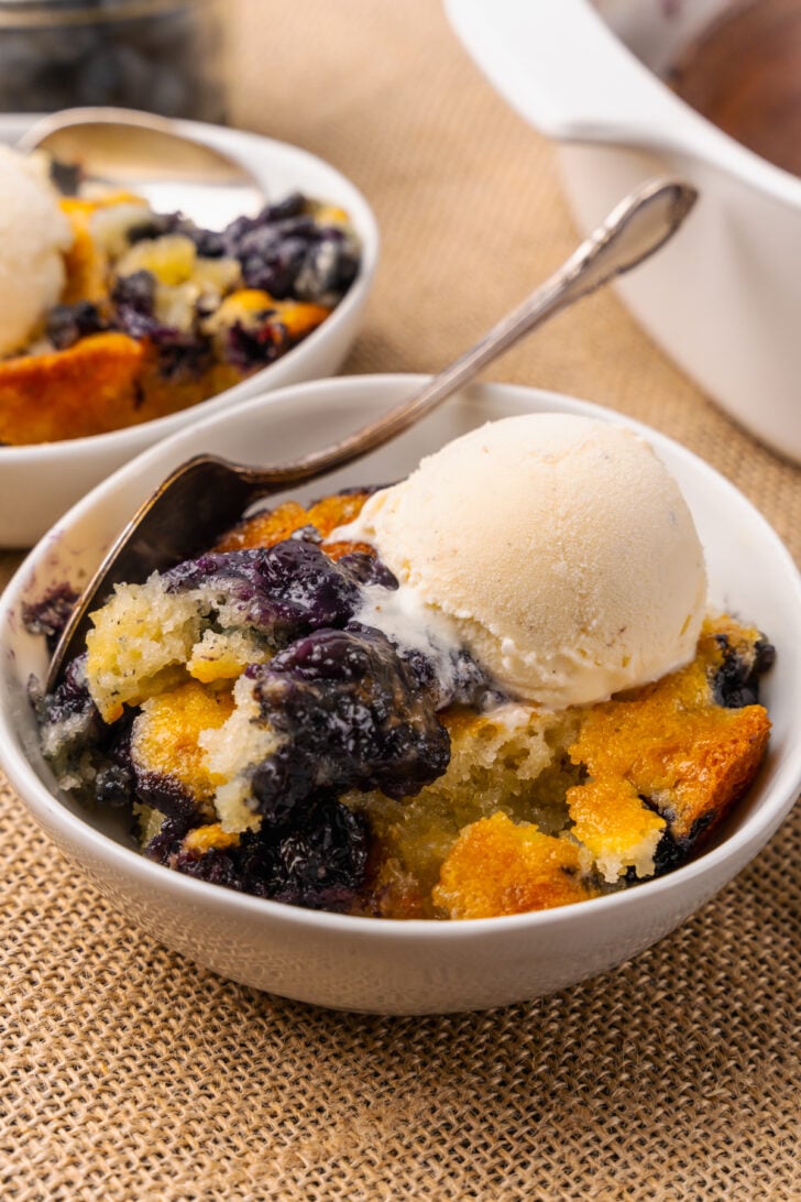 White bowl with blueberry cobbler and a scoop of vanilla ice cream resting on a tan placemat with silver forks.