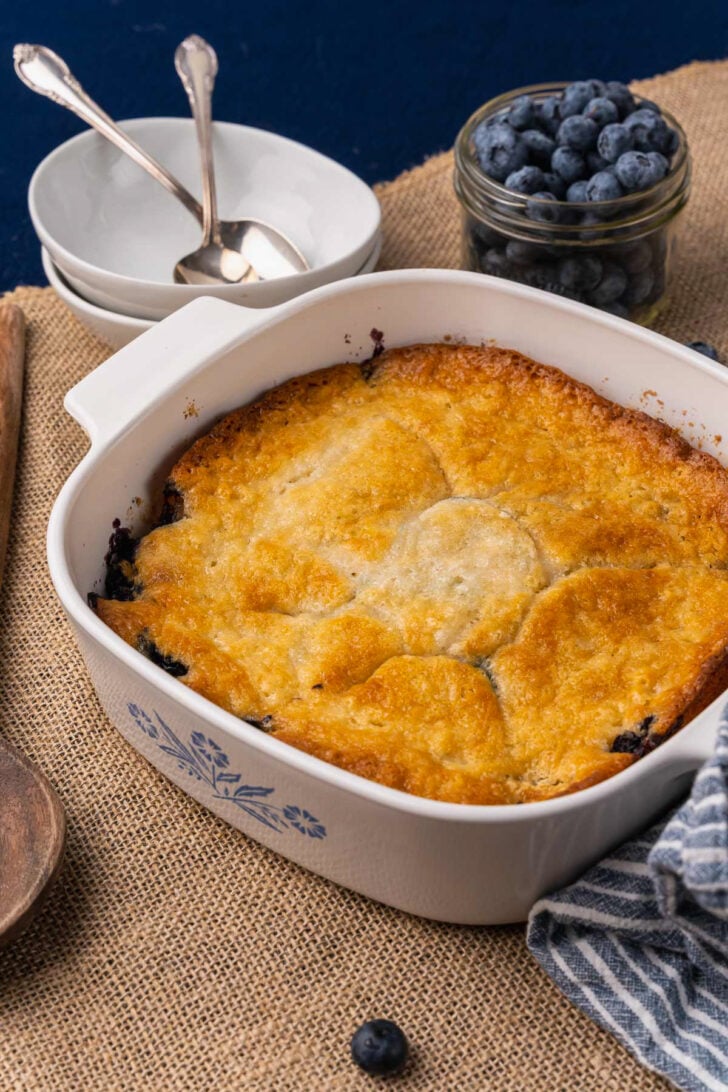 Baked blueberry cobbler in a white casserole dish resting on a tan placemat.