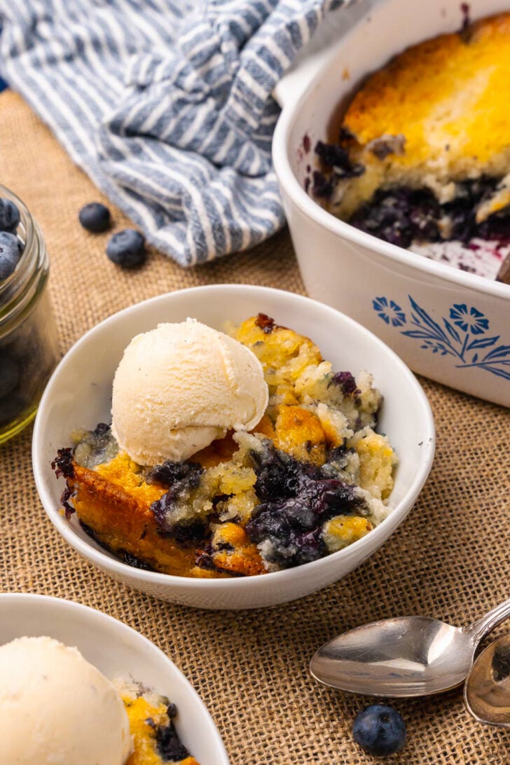 White bowl with blueberry cobbler and a scoop of vanilla ice cream resting on a tan placemat with silver forks.