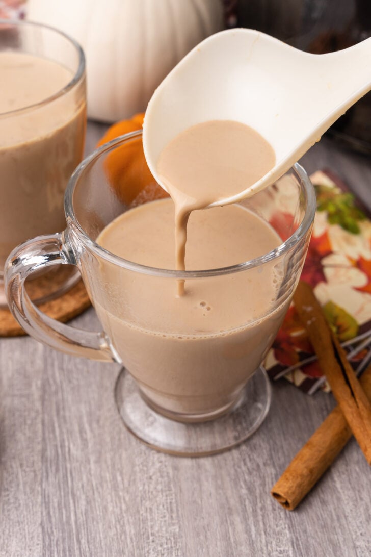 White ladle spooning in pumpkin spice latte into a glass mug sitting on a wooden table.