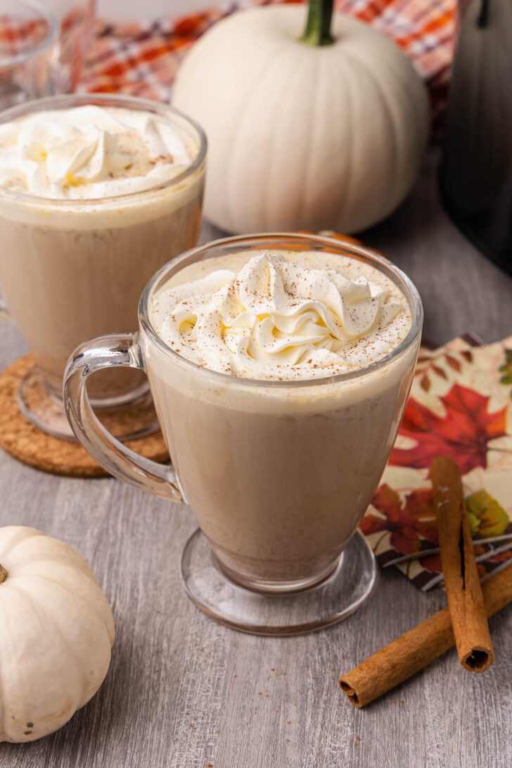 Creamy pumpkin spice latte topped with whipped cream in glass mugs on a wood table with cinnamon sticks.