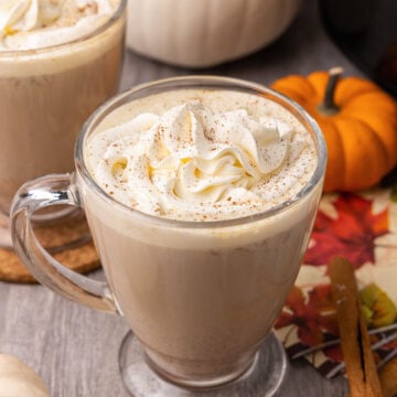 Creamy pumpkin spice latte topped with whipped cream in glass mugs on a wood table with cinnamon sticks.