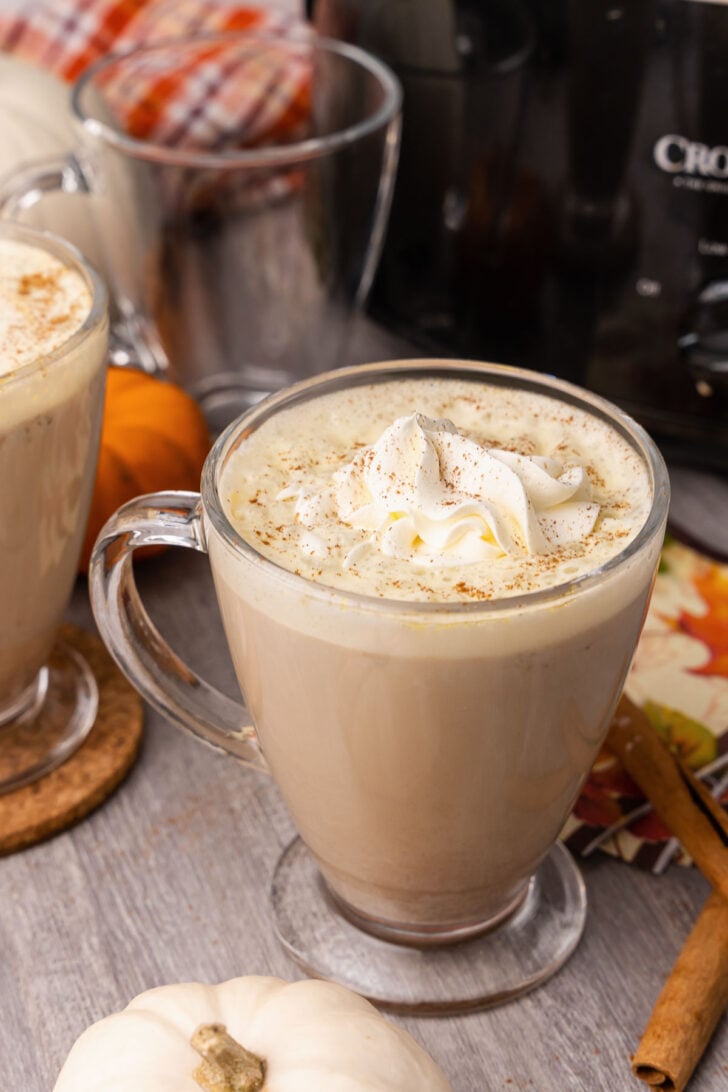Creamy pumpkin spice latte topped with whipped cream in glass mugs on a wood table with cinnamon sticks.