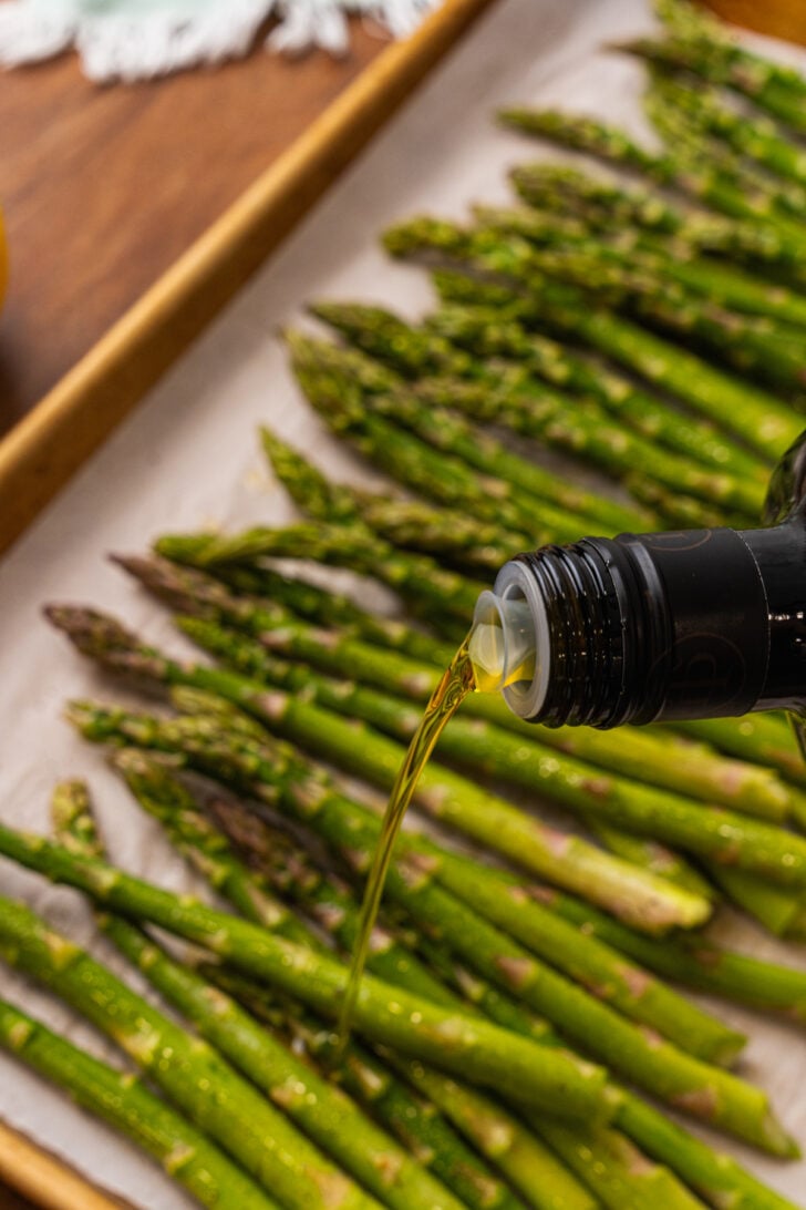 Olive oil drizzled over asparagus spears on a parchment lined baking sheet.