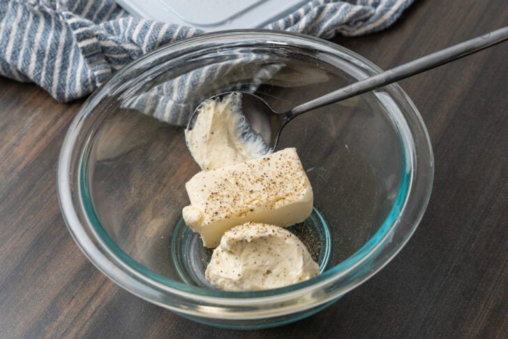 Clear glass board with cream cheese and herb cheese and black pepper with a black mixing spoon resting on a black cutting board.
