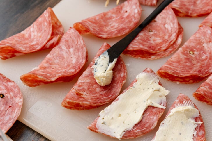 Salami slices folded in half spread with herb, cream cheese spread with a black knife on a cutting board.