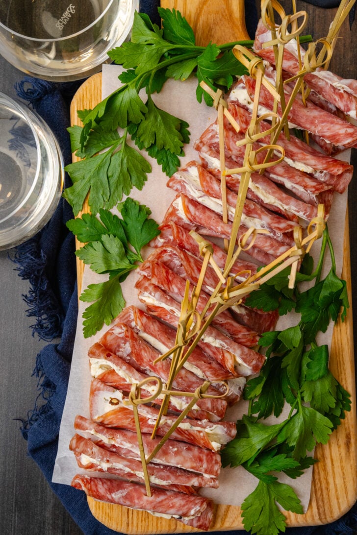Wooden cutting board with cream cheese filled salami slices with a wooden pick garnished with parsley and served with white wine in glasses.