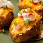 Stuffed baked potatoes with bacon, and cheese then topped with more bacon bits, sour cream and green onion slices on a green plate.