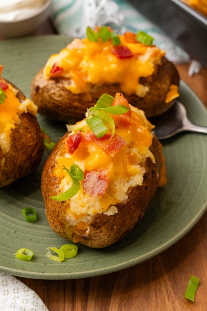 Stuffed baked potatoes with bacon, and cheese then topped with more bacon bits, and green onion slices on a green plate.