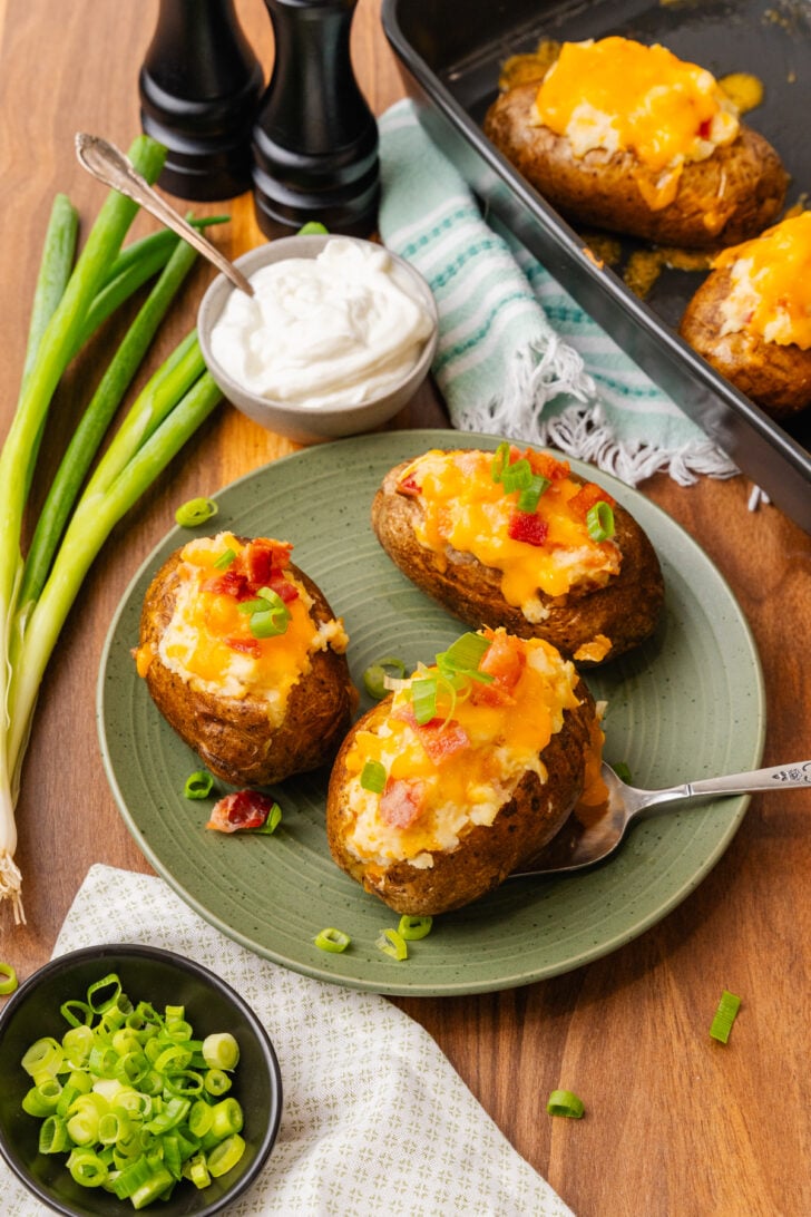 Stuffed baked potatoes with bacon, and cheese then topped with more bacon bits, sour cream and green onion slices on a green plate.