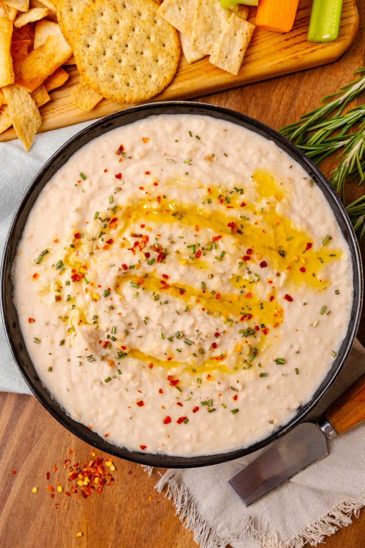 Black bowl with white bean dip topped with red pepper flakes, dried chives, and red pepper flakes, on a wooden board served with crackers.