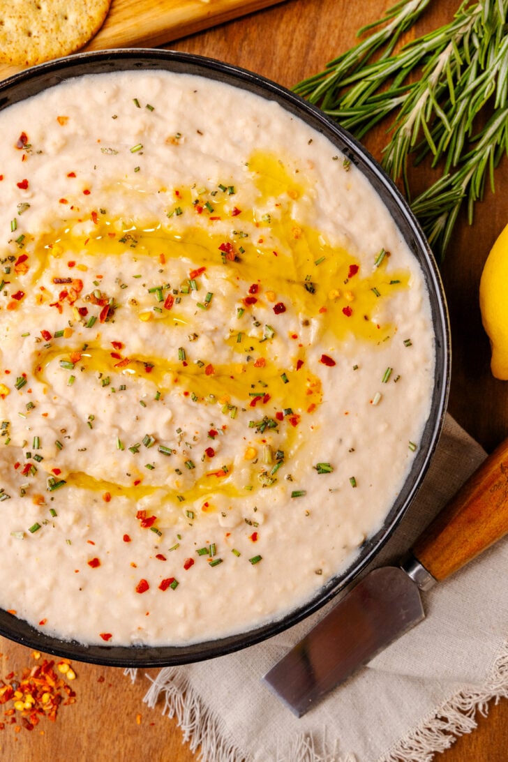 Black bowl with white bean dip topped with red pepper flakes, dried chives, and red pepper flakes, on a wooden board.
