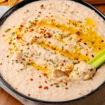 Black bowl with white bean dip topped with red pepper flakes, dried chives, and red pepper flakes, dipped with a celery stick.