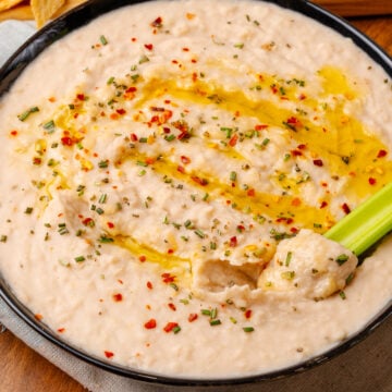 Black bowl with white bean dip topped with red pepper flakes, dried chives, and red pepper flakes, dipped with a celery stick.