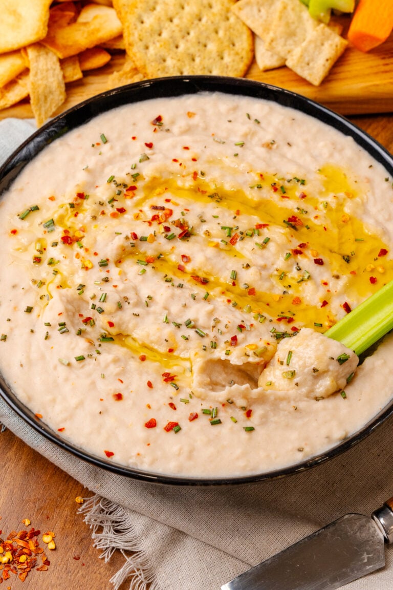 Black bowl with white bean dip topped with red pepper flakes, dried chives, and red pepper flakes, dipped with a celery stick.