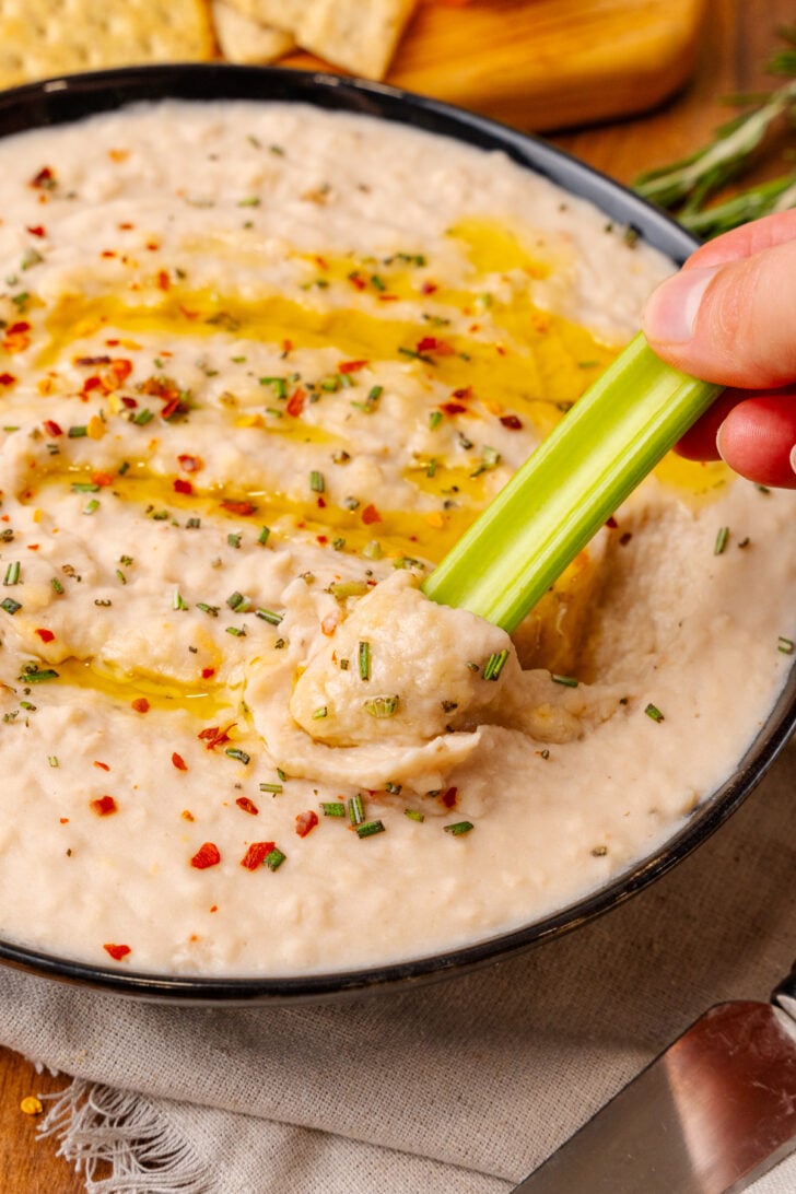 Black bowl with white bean dip topped with red pepper flakes, dried chives, and red pepper flakes, dipped with a celery stick.