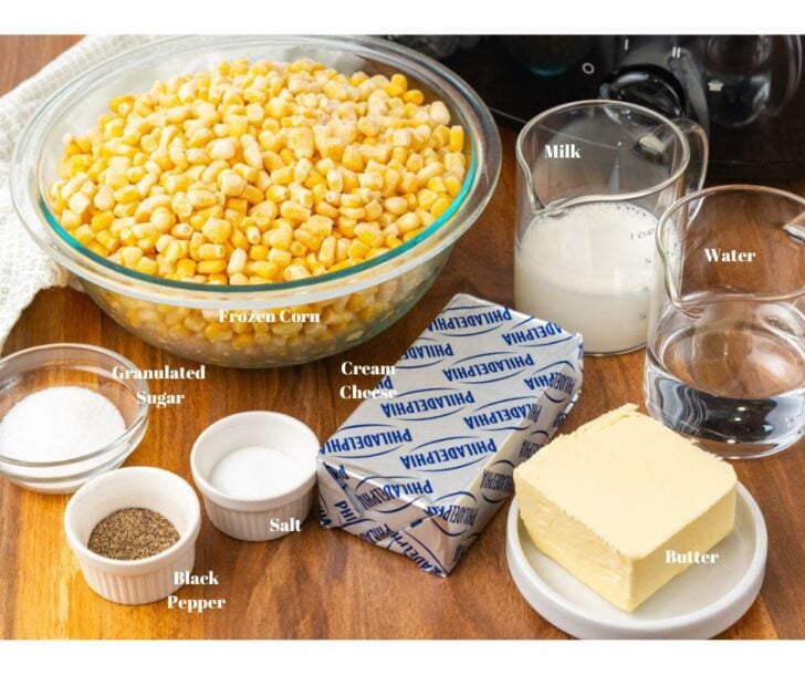 Frozen corn, milk, water, butter, cream cheese, salt, pepper and sugar in glass bowls on a brown counter top.