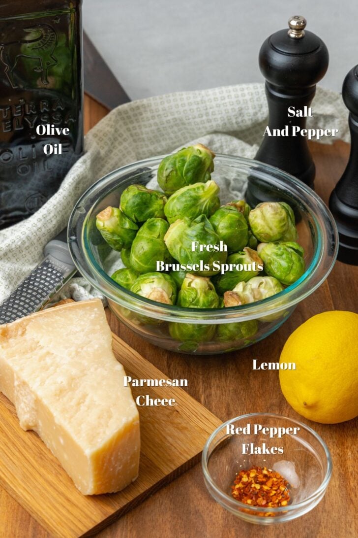Olive oil, salt and pepper in black, fresh brussels sprouts, block of parmesan cheese, whole fresh lemon, red pepper flakes on a cutting board.