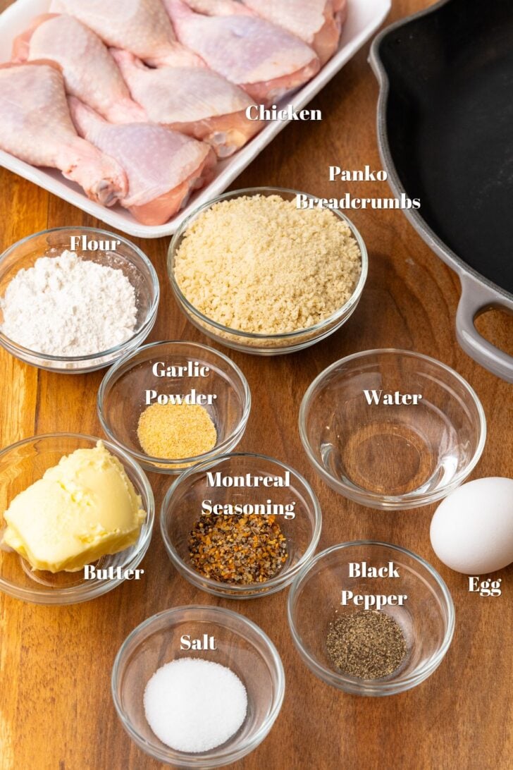 Chicken legs, panko breadcrumbs, flour, garlic powder, water, butter, montreal seasoning, egg, pepper and salt in glass bowls on a wooden table.