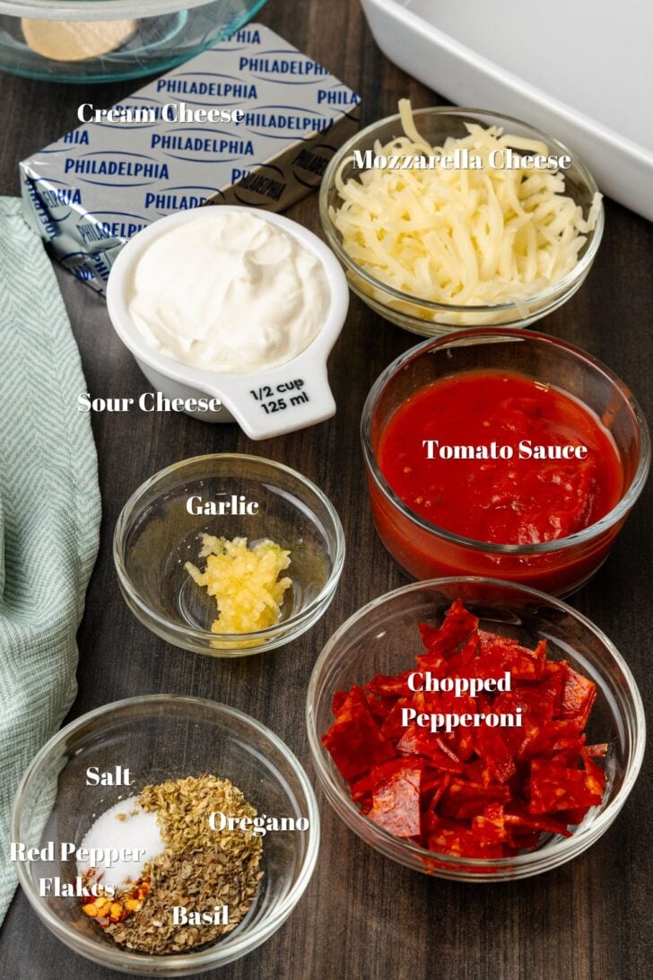 Cream cheese, mozzarella cheese, sour cream, tomato saue, garlic, chopped pepperoni salt, red pepper, oregano and basil on a wooden counter.
