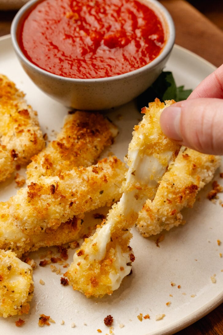 Hand holding golden-fried cheese sticks 9 with crispy coating, served on white plate with red dipping sauce in bowl