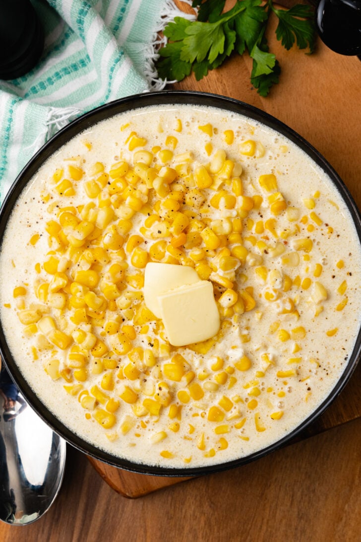 Crock Pot Cream Corn served in a black bowl topped with butter on a wooden board.