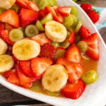 Strawberroes, bananas and green grapes in a dressing served in a white oblong bowl.