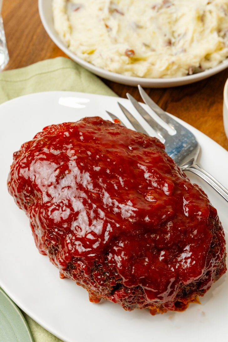 Whole cooked meatloaf on a white plate with a silve fork for serving.