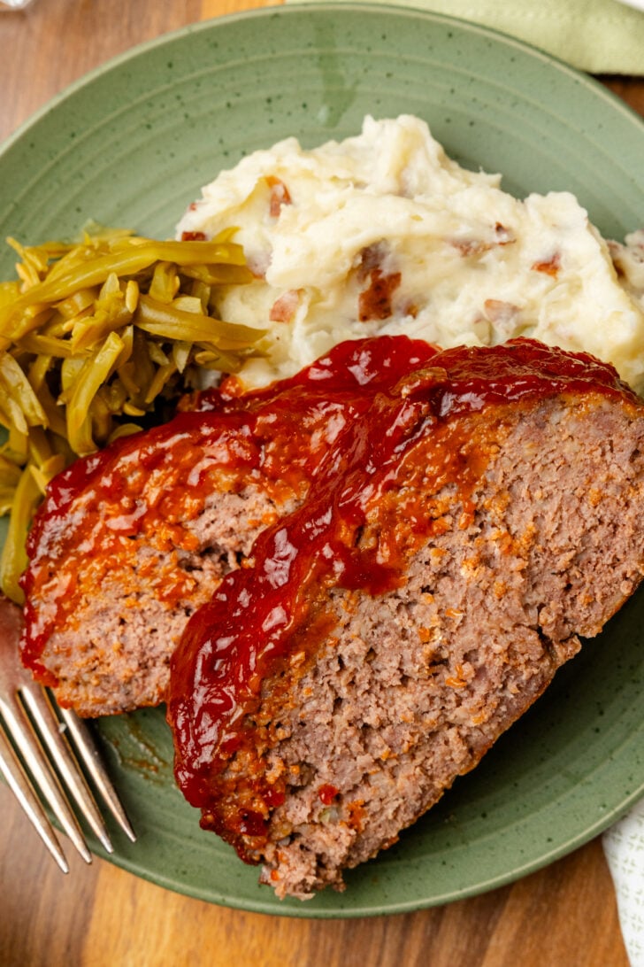 Sliced of meatloaf topped with a sweet glaze served with mashed potatoes and green beans on a green plate.