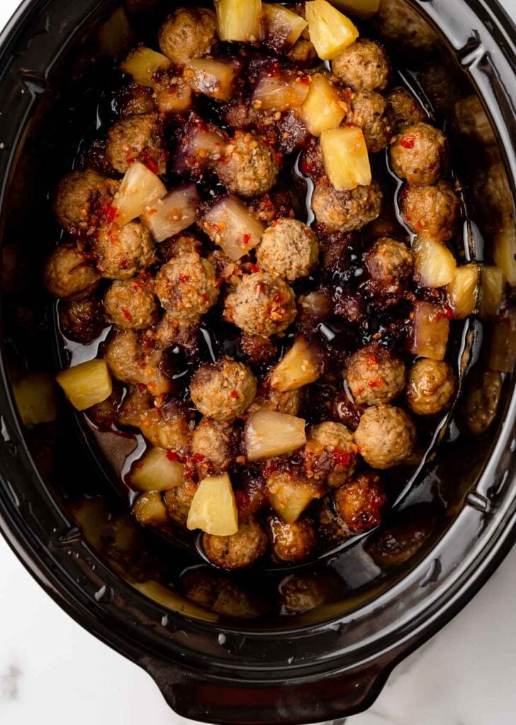 Meatballs, pineapple chunks, sauce, and red pepper seasoning in a black crock pot.