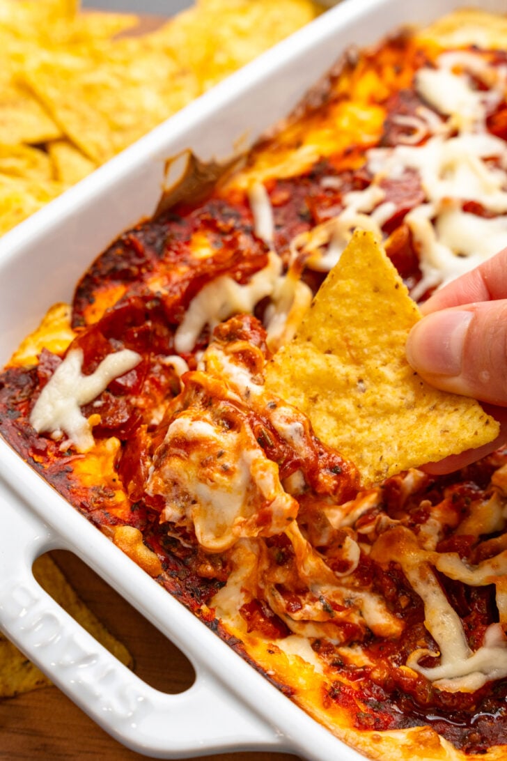 Hand holding a tortilla chip with a scoop of Pepperonit dip with tomato sauce and cheese.