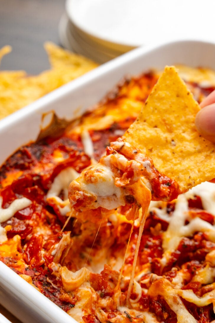 Hand holding a tortilla chip with a scoop of Pepperonit dip with tomato sauce and cheese.