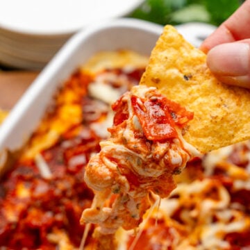 Hand holding a tortilla chip with a scoop of Pepperonit dip with tomato sauce and cheese.