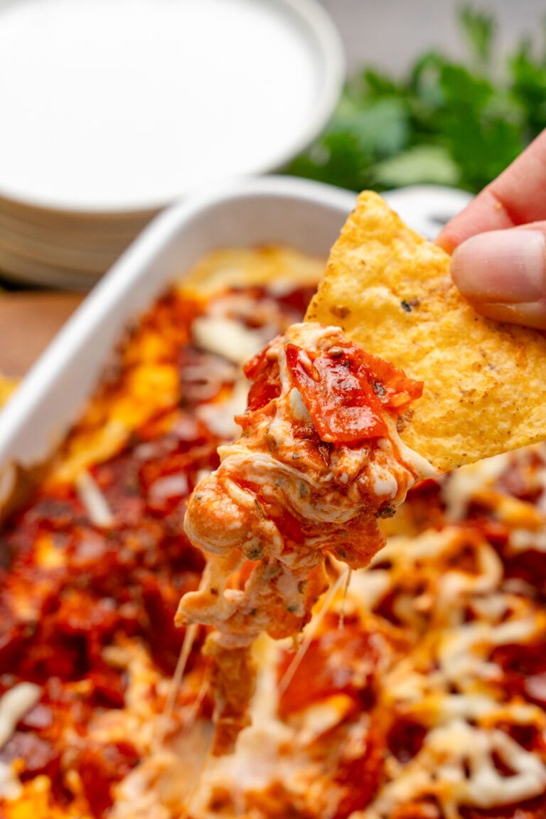 Hand holding a tortilla chip with a scoop of Pepperonit dip with tomato sauce and cheese.